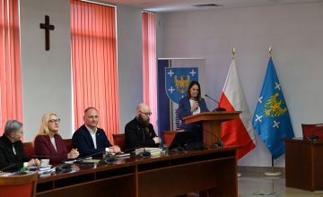 Sala konferencyjna, przy mównicy stoi osoba, z lewej strony siedzą trzy osoby. W tle flaga Polski i Powiatu